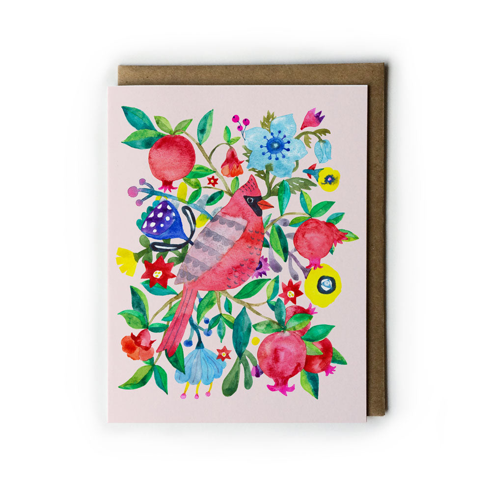 Colorful watercolor greeting card featuring a red cardinal bird surrounded by vibrant flowers, pomegranates, and festive foliage on a light pink background, paired with a kraft envelope for a charming holiday design