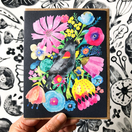 Vibrant watercolor greeting card featuring a blackbird surrounded by colorful flowers on a black background, held against a patterned surface