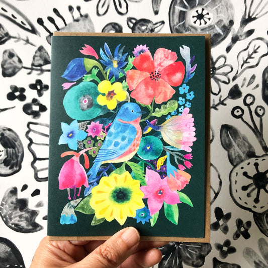 Vibrant watercolor greeting card featuring a colorful bluebird surrounded by diverse flowers and foliage on a dark green background, held in hand against a black and white floral pattern