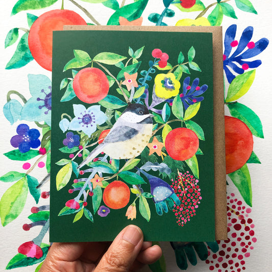 Watercolor holiday greeting card featuring a chickadee bird perched among vibrant oranges, colorful flowers, and lush greenery on a dark green background, held by a hand against a floral patterned surface