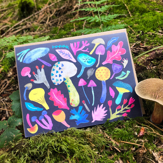 Colorful mushroom greeting card displayed in forest setting, featuring vibrant watercolor illustrations of various mushroom species and foliage on dark background, surrounded by real moss and fungi
