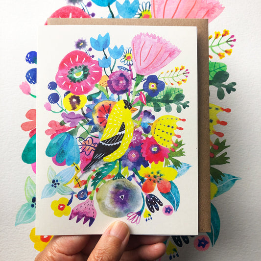 Vibrant watercolor greeting card featuring a yellow goldfinch perched among colorful flowers, including pink, blue, and purple blooms, with a kraft envelope shown beneath. Hand holding the card displays its size and texture.