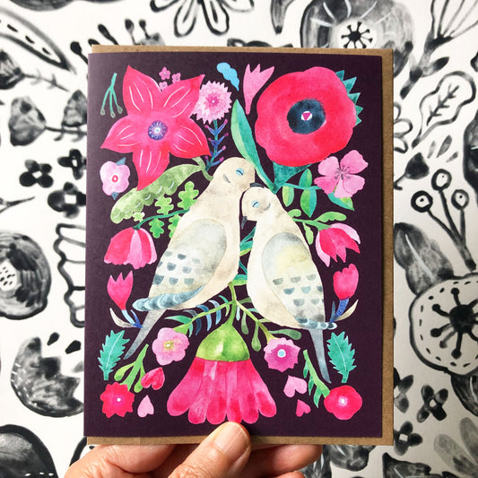 Colorful greeting card featuring two mourning doves surrounded by vibrant watercolor flowers and foliage, including pink poppies and tulips, on a dark background. Hand holding the card against a black and white patterned surface.