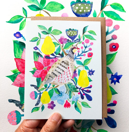 Watercolor greeting card featuring a partridge surrounded by vibrant pears, flowers, and foliage. The colorful design includes red, blue, yellow, and green elements, displayed against a kraft paper envelope background.