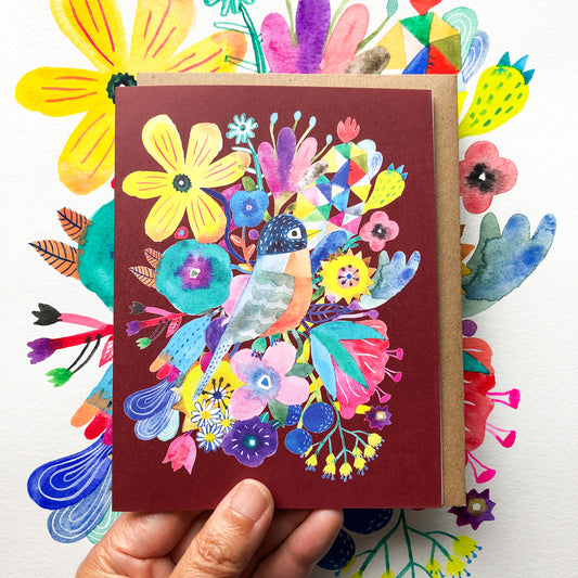 Colorful greeting card featuring a vibrant watercolor illustration of a robin surrounded by various flowers and foliage on a burgundy background, held against a floral patterned surface