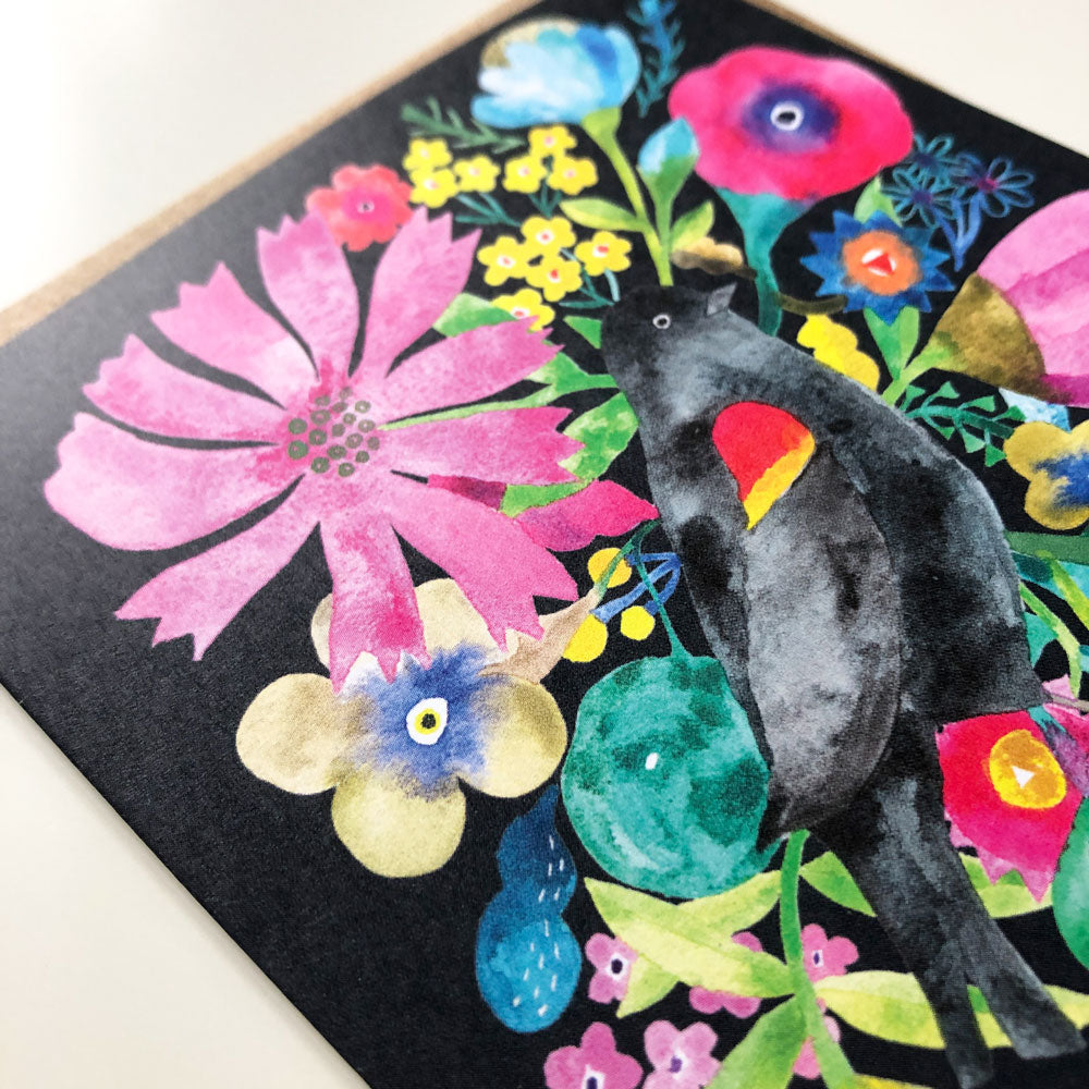 Vibrant watercolor greeting card featuring a black bird perched among colorful flowers, including a large pink bloom, on a dark background. The card displays a variety of floral shapes and hues, creating a lively and artistic composition.