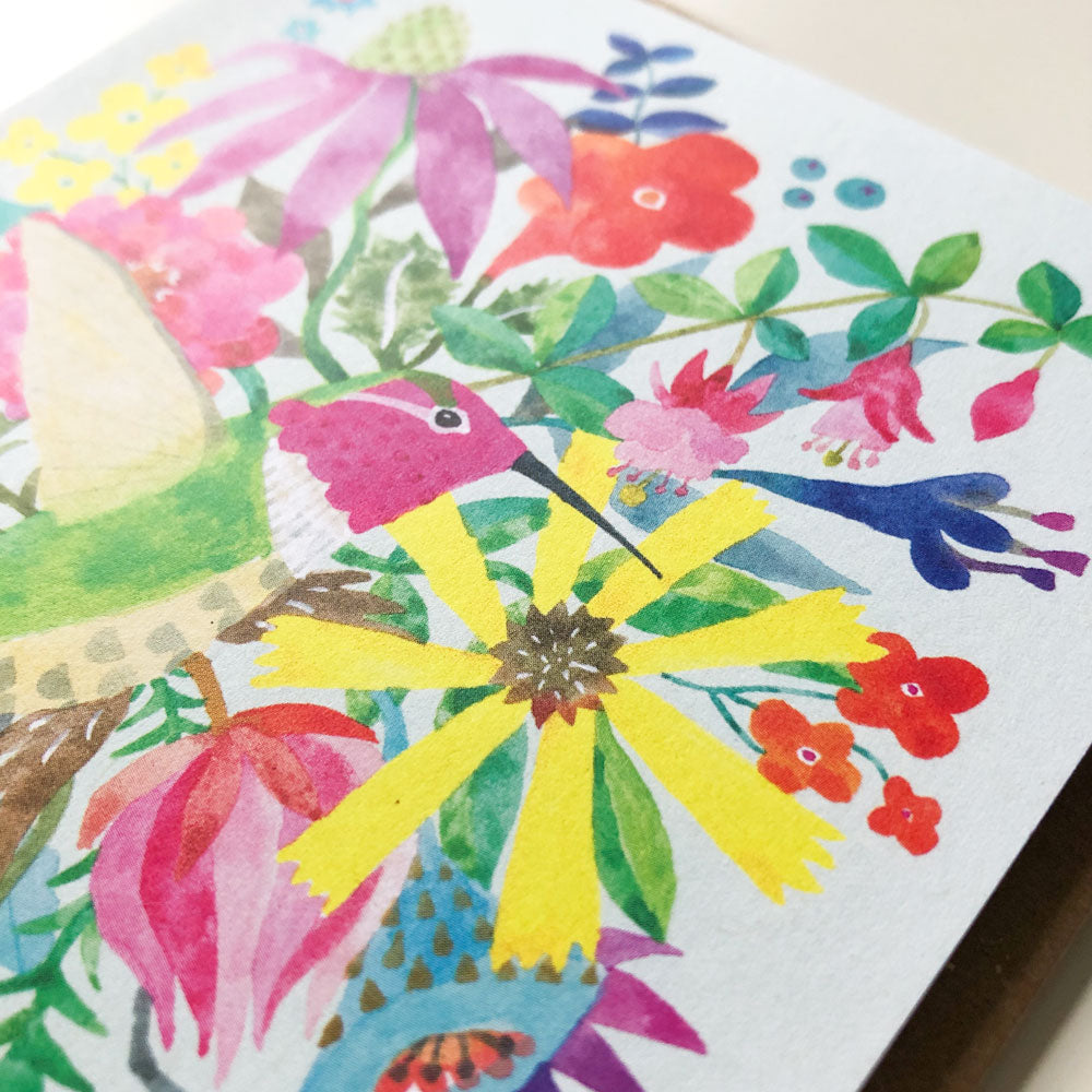 Colorful watercolor greeting card featuring a vibrant hummingbird surrounded by various flowers and foliage, including yellow, pink, red, and blue blooms, on white paper