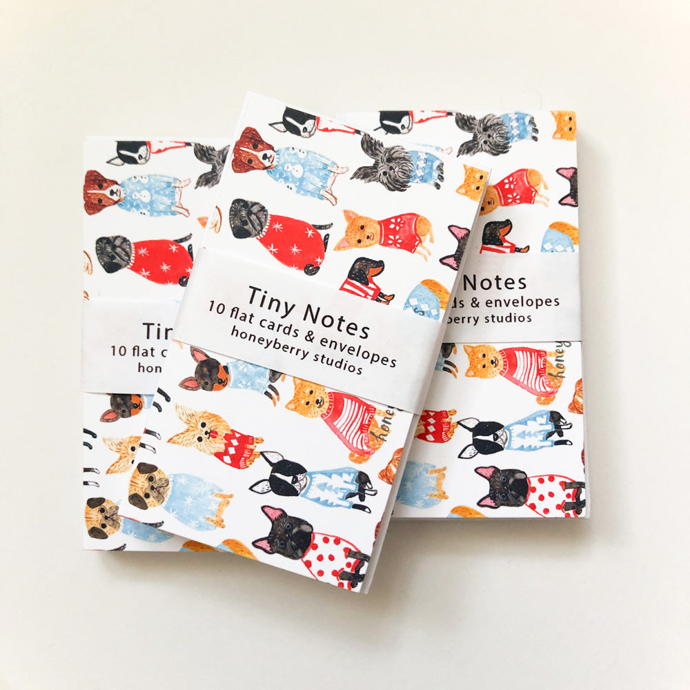 Two sets of tiny note cards with colorful illustrations of various dog breeds wearing sweaters and festive outfits. The cards are bundled with white bands labeled 'Tiny Notes' and '10 flat cards & envelopes'.