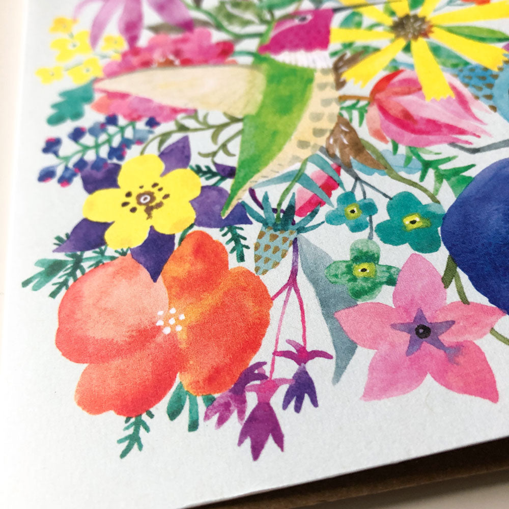 Vibrant watercolor illustration of colorful flowers and a hummingbird, featuring various blooms in shades of red, yellow, purple, and pink with green foliage, perfect for a greeting card design