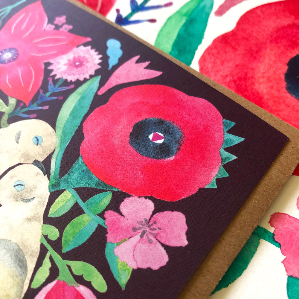 Vibrant watercolor greeting card featuring a large red poppy flower, pink blossoms, and green leaves on a dark background, with kraft envelope visible