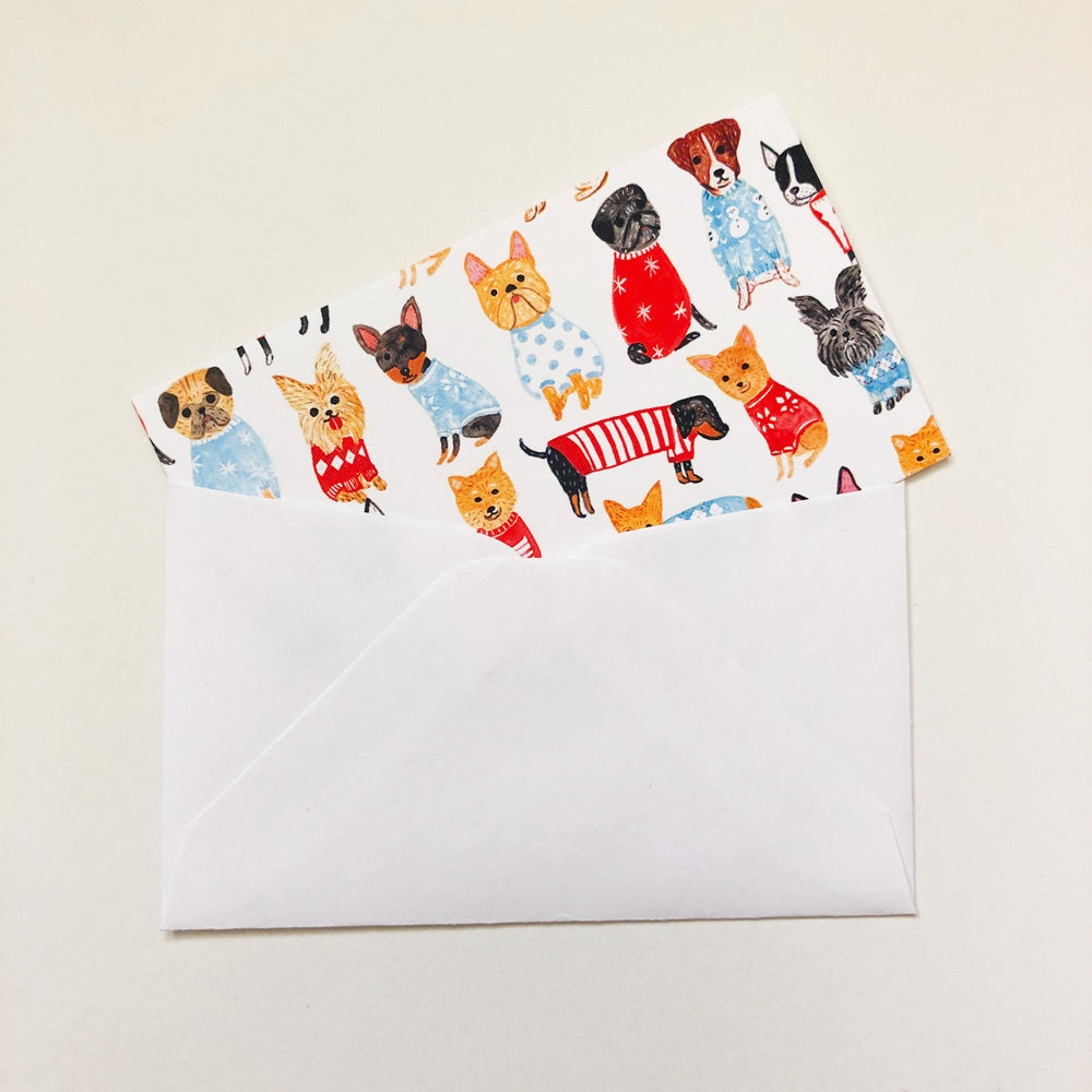 Tiny note card featuring colorful illustrations of various dog breeds wearing sweaters, partially inserted into a white envelope, displayed on a light background