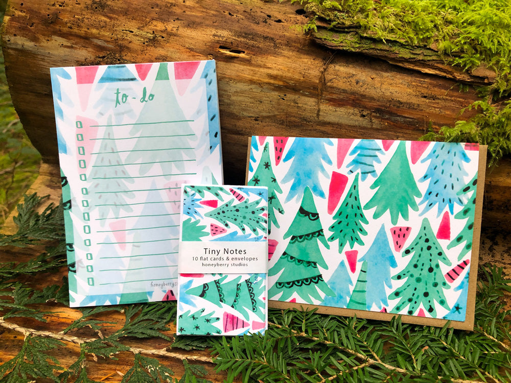 Christmas Forest To-Do List notepad, greeting card, and tiny notes displayed on wooden log and pine branches, featuring colorful watercolor illustrations of Christmas trees in green, blue, and pink