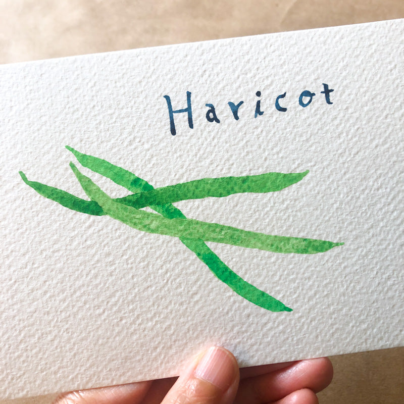 Watercolor painting on textured paper featuring the word "Haricot" in blue script above a green illustration of string beans, held by fingertips at the bottom edge