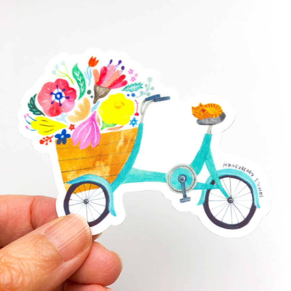 Colorful vinyl sticker featuring a teal bicycle with a basket full of vibrant flowers and a ginger cat riding on the seat, held by fingers against a white background