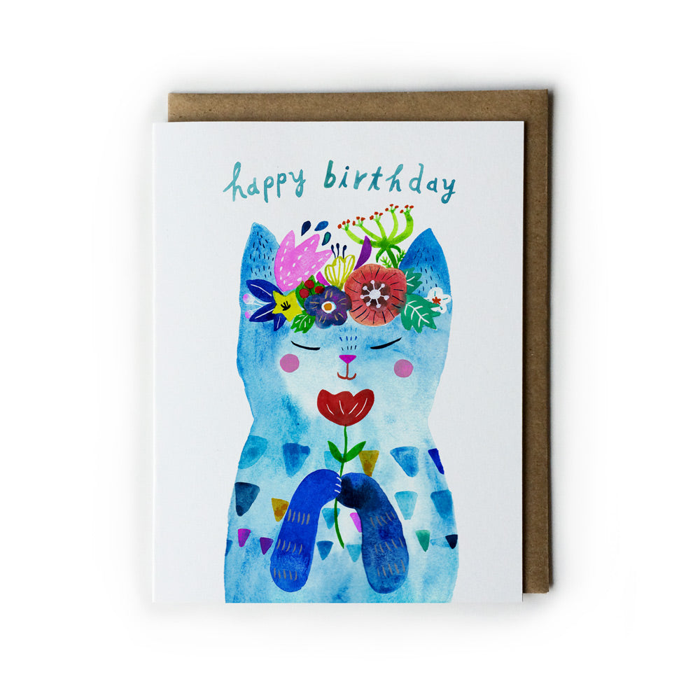 Colorful watercolor birthday card featuring a whimsical blue cat with floral crown, holding a red flower, against white background with kraft paper envelope. Text reads "happy birthday" in teal script.
