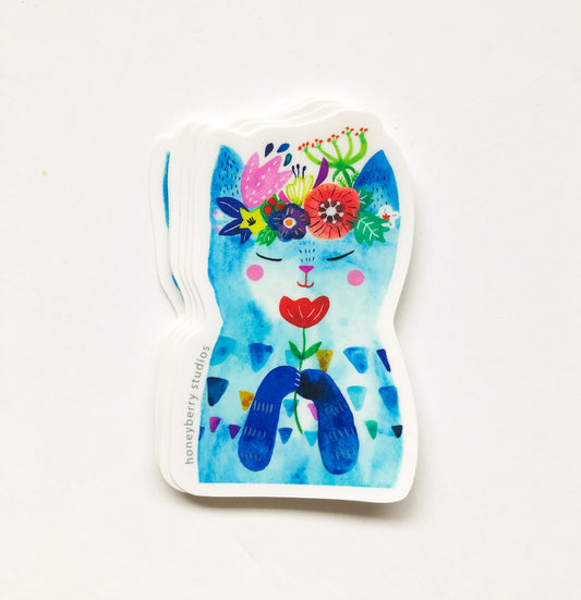 Whimsical blue cat-shaped sticker with colorful floral crown, rosy cheeks, and red flower on its body, featuring a playful and artistic design on white background