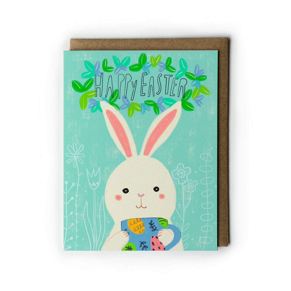 Cute Easter greeting card featuring a cartoon bunny holding a colorful mug, with "Happy Easter" text surrounded by green leaves on a mint background, accompanied by a kraft envelope