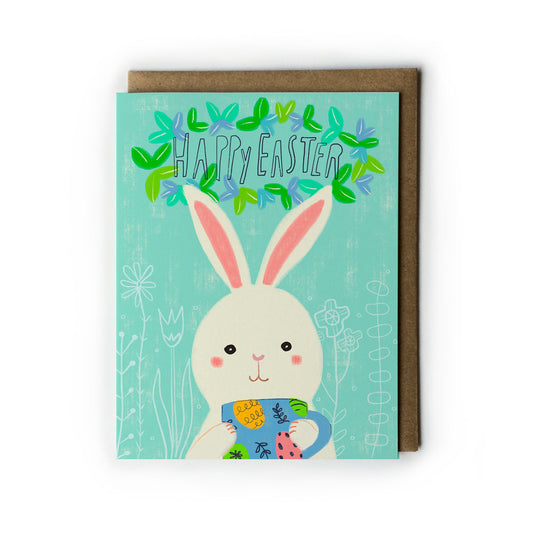 Cute Easter greeting card featuring a cartoon bunny holding a colorful mug, with "Happy Easter" text surrounded by green leaves on a mint background, accompanied by a kraft envelope