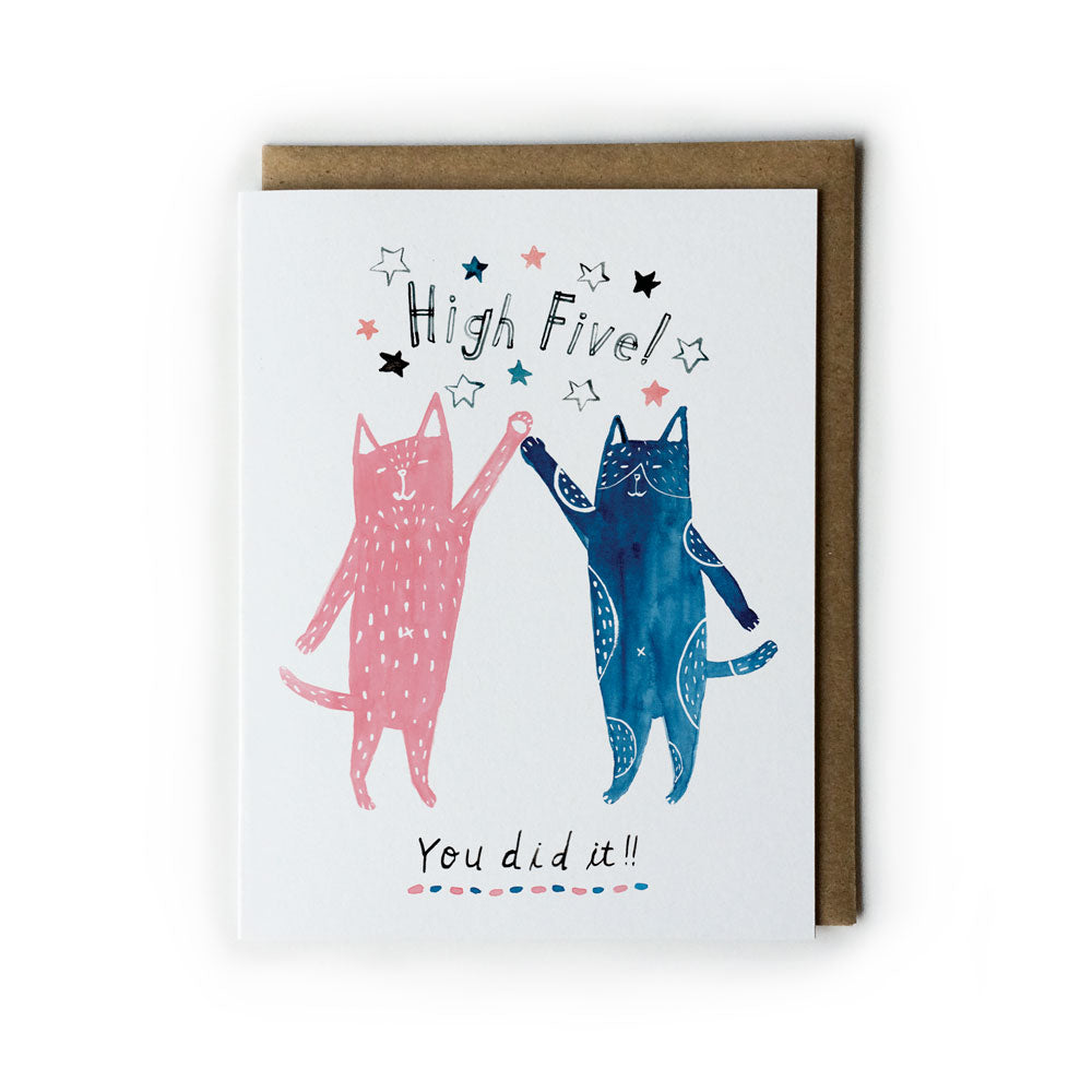 Watercolor greeting card featuring two cartoon cats, one pink and one navy blue, giving a high five with "High Five!" and "You did it!" text, surrounded by stars, on white card with kraft envelope