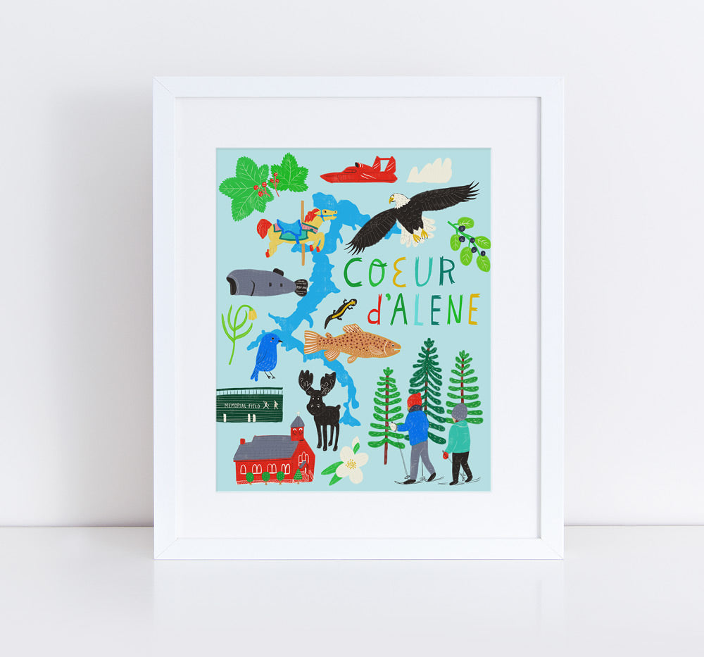Colorful illustration of Coeur d'Alene, Idaho featuring local landmarks, wildlife, and activities. Includes hand-lettered text, lake, trees, eagle, fish, plane, and people enjoying outdoor pursuits. Framed art print on white background.