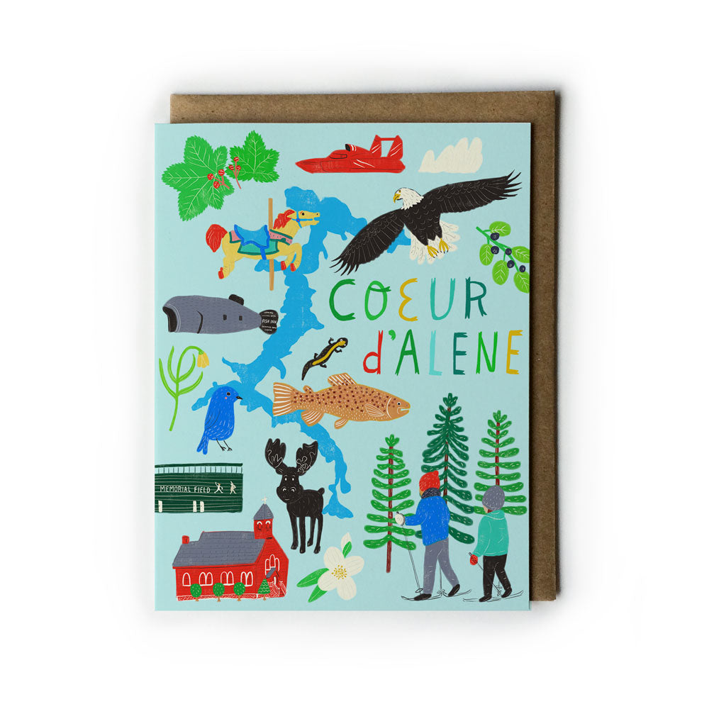 Colorful illustrated greeting card featuring Coeur d'Alene landmarks, wildlife, and activities. Includes lake, fish, eagle, deer, trees, hikers, and hand-lettered text. Card shown with brown envelope on white background.