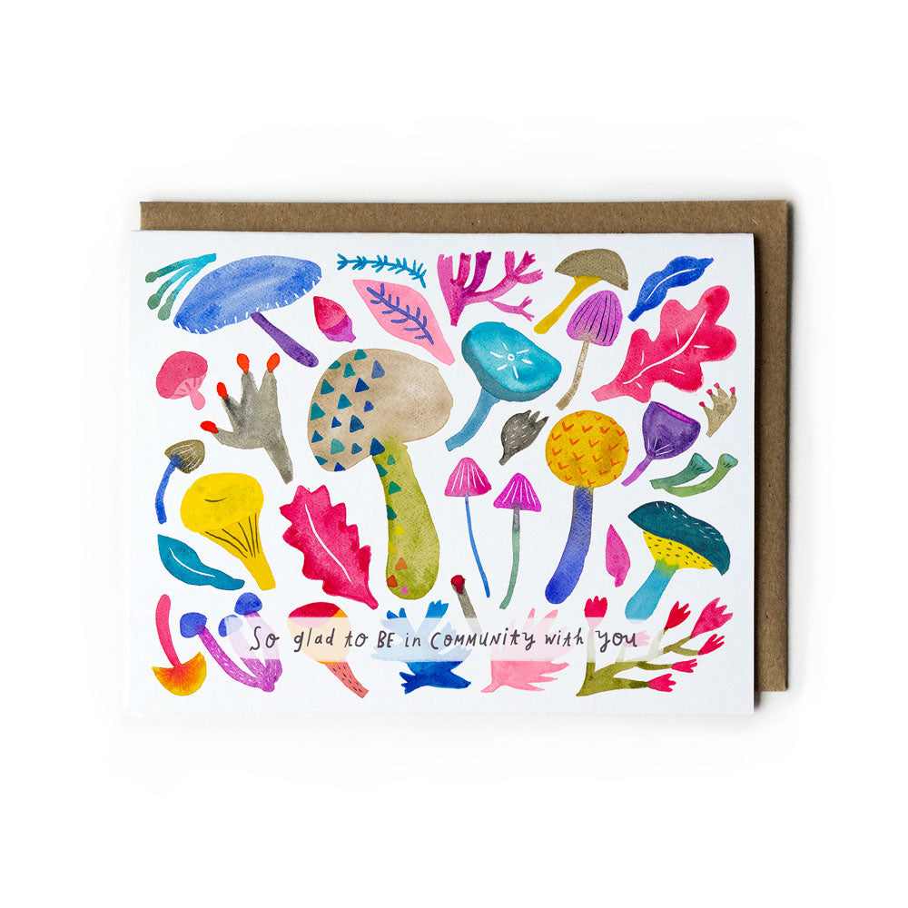 Colorful watercolor greeting card featuring diverse mushrooms, leaves, and abstract shapes with text "So glad to be in community with you" in whimsical handwritten style, accompanied by kraft paper envelope.