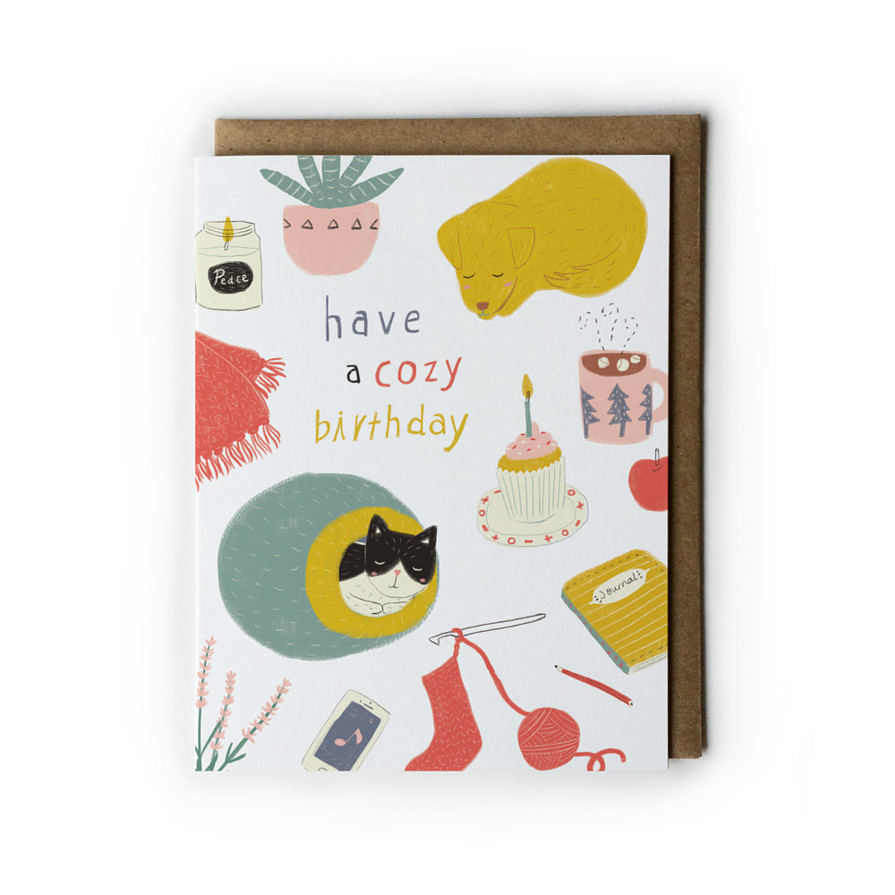Birthday card featuring cozy elements: sleeping cat and dog, cupcake, knitting supplies, plant, candle, and text "have a cozy birthday" on white background with brown envelope