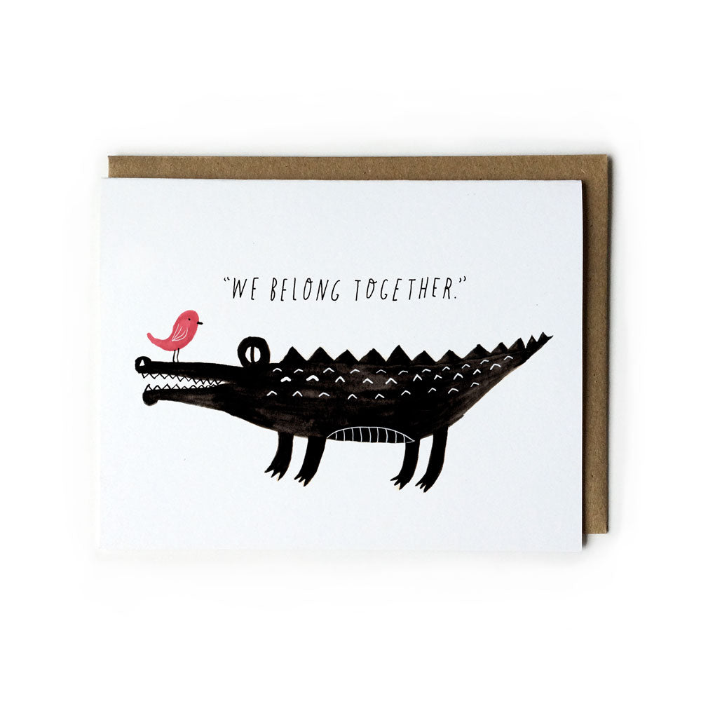Watercolor greeting card featuring a black crocodile with a pink bird perched on its nose, with the text "We Belong Together" above, accompanied by a kraft envelope