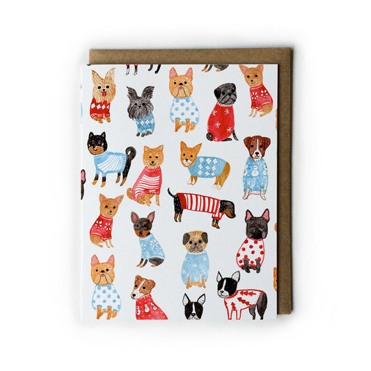 Holiday greeting card featuring various small dog breeds wearing colorful sweaters and festive outfits, arranged on a white background with a kraft paper envelope