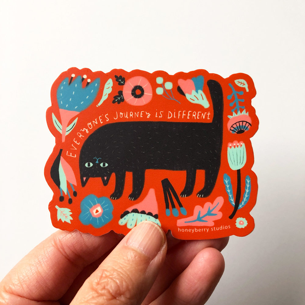 Colorful vinyl sticker featuring a black cat surrounded by floral designs and text "Everyone's journey is different" in a vibrant orange frame, held by fingertips to show size and material