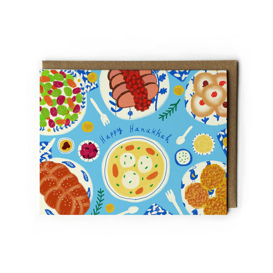 Colorful Hanukkah greeting card featuring illustrated traditional foods on blue background, including matzo ball soup, latkes, challah bread, and sufganiyot. Text reads "Happy Hanukkah" in center. Kraft paper envelope included.