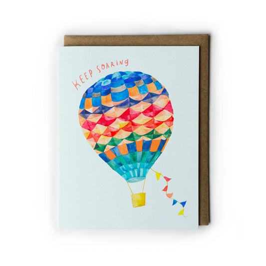 Colorful hot air balloon greeting card with "Keep Soaring" text, featuring geometric patterns and vibrant hues on light blue background, accompanied by kraft paper envelope