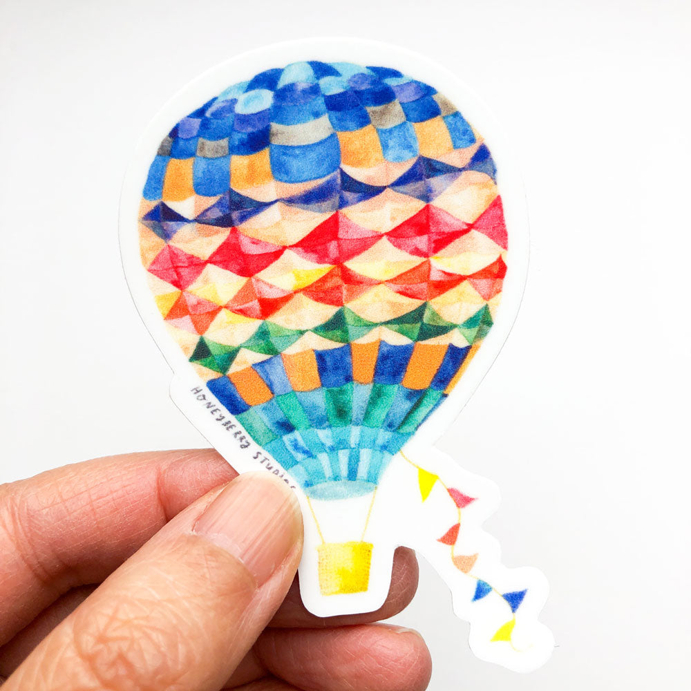 Colorful hot air balloon vinyl sticker with geometric pattern design, featuring vibrant blues, reds, yellows, and greens, held between fingers to showcase its size and detail