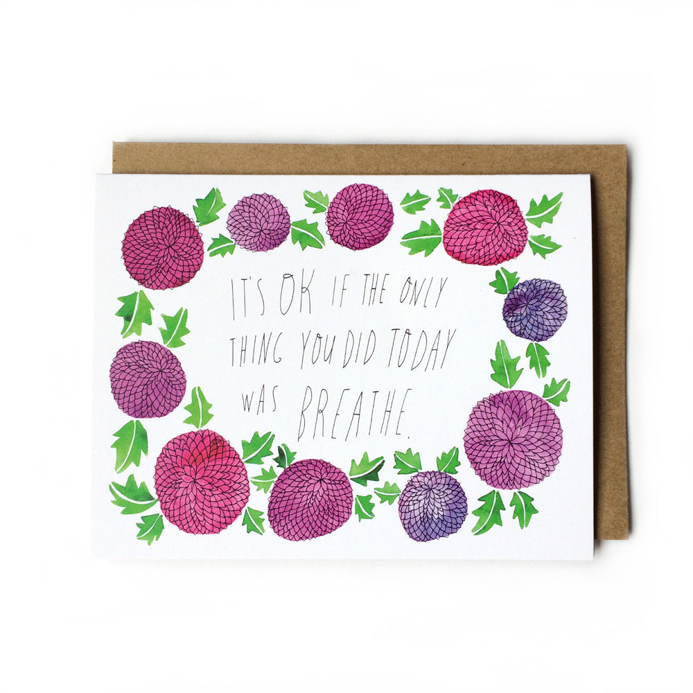 Greeting card with hand-drawn floral border featuring purple and pink flowers, green leaves, and handwritten text "It's OK if the only thing you did today was breathe" on white background, paired with kraft paper envelope