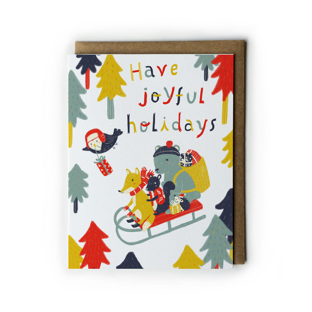 Colorful holiday card featuring woodland animals on a sled, surrounded by festive trees, with "Have joyful holidays!" text in bright letters, accompanied by a kraft paper envelope