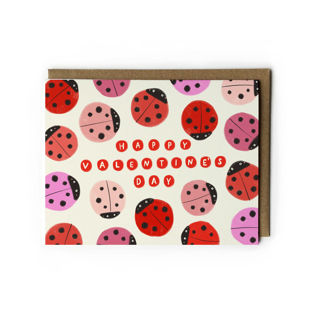 Whimsical Valentine's Day card featuring colorful ladybug illustrations in red and pink on white background, with hand-lettered "Happy Valentine's Day" message. Paired with kraft paper envelope.
