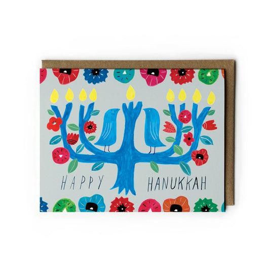 Colorful Hanukkah card featuring a stylized blue menorah tree with birds, flowers, and candles. Handwritten "Happy Hanukkah" message. Vibrant floral border. Kraft paper envelope included.