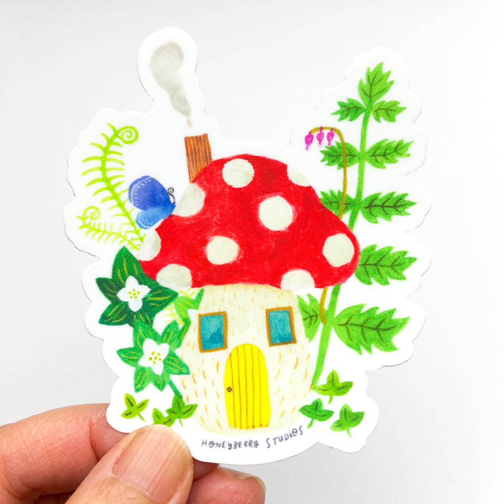 Whimsical vinyl sticker featuring a red and white polka dot mushroom house with yellow door, surrounded by colorful plants and flowers, held by fingers to show scale
