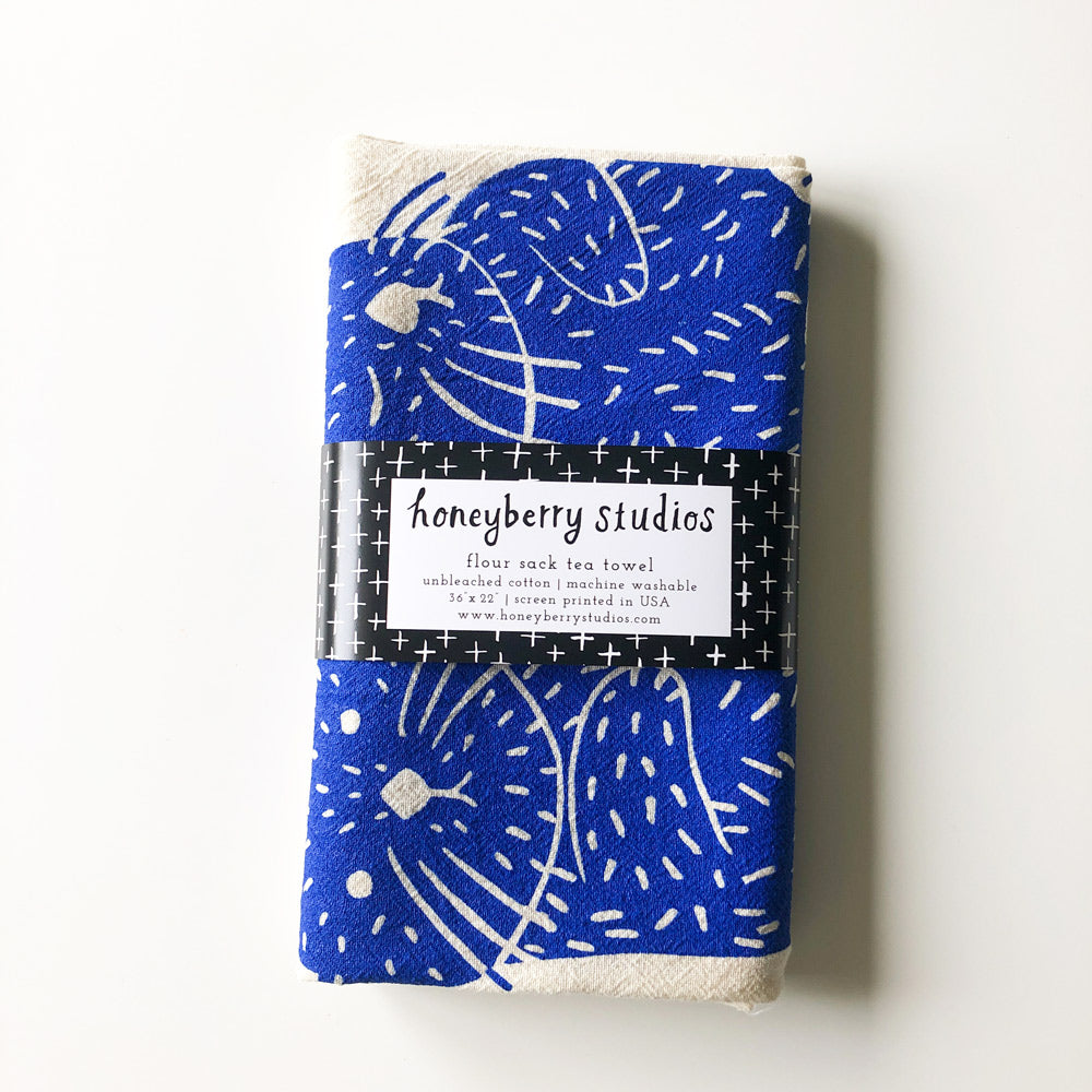 Flour sack tea towel with vibrant blue floral pattern, folded and packaged with Honeyberry Studios label, showcasing handmade quality and artistic design for kitchen use