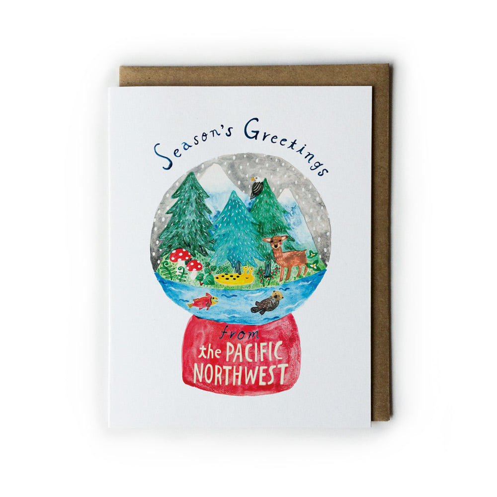 White greeting card featuring a watercolor illustration of a snow globe with Pacific Northwest scenery, including evergreen trees, mountains, wildlife, and water, on a red base labeled "The Pacific Northwest". Accompanied by a kraft paper envelope.