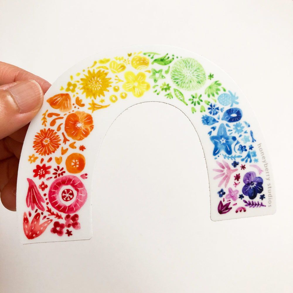 Colorful rainbow arch sticker featuring diverse flowers and botanical elements arranged in a spectrum from red to purple, held by fingers against a white background