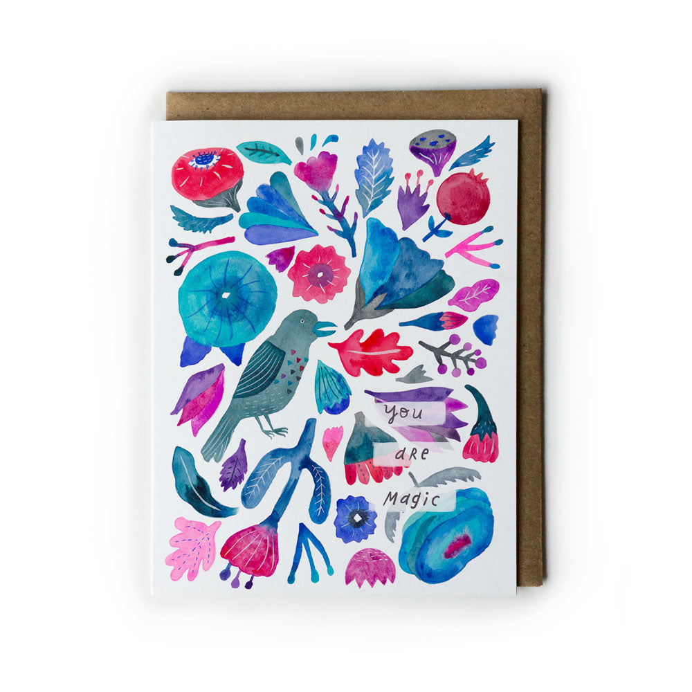 Colorful watercolor greeting card featuring a raven among vibrant flowers, leaves, and the words "you are magic" in a whimsical folk art style, paired with a kraft paper envelope.