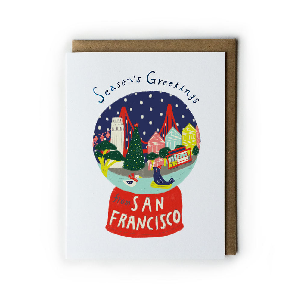 Season's Greetings card featuring a San Francisco snow globe with colorful houses, a cable car, Christmas tree, and iconic landmarks against a snowy night sky, alongside a kraft paper envelope