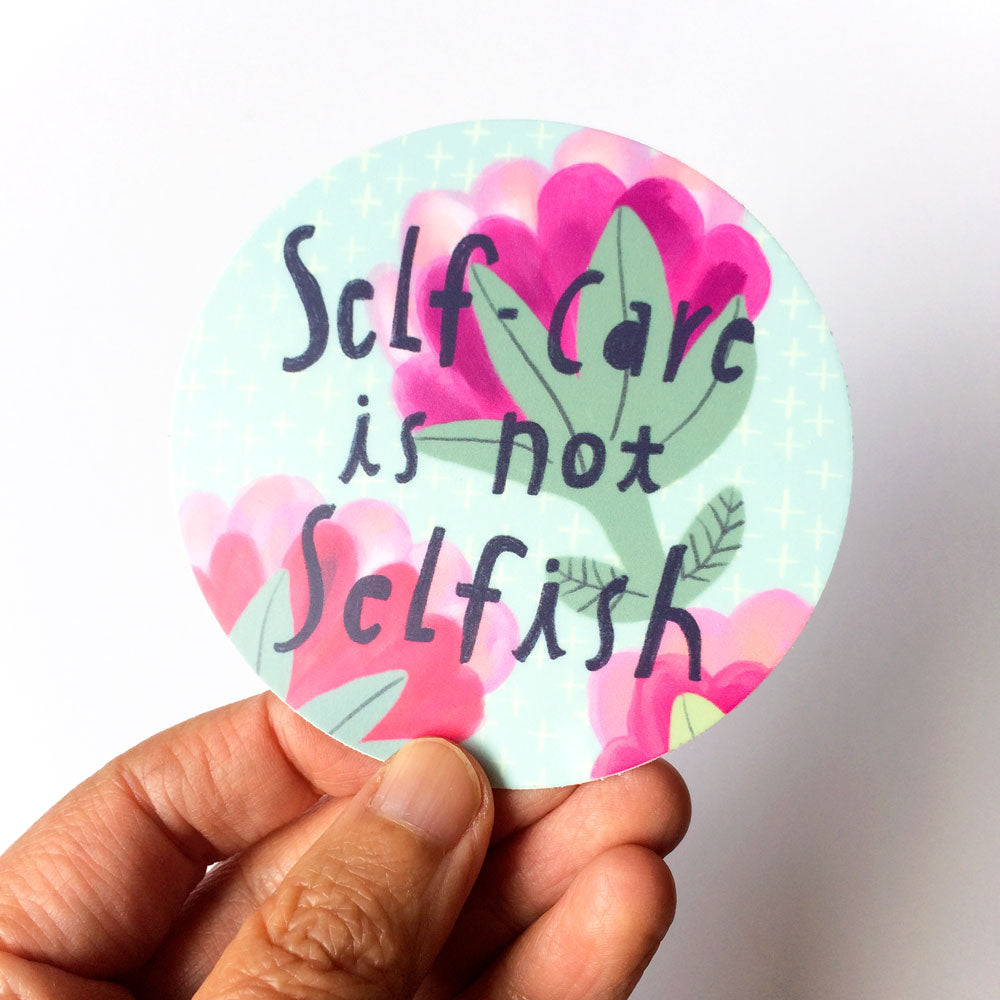 Circular vinyl sticker with 'Self-care is not selfish' text, surrounded by pink heart shapes and green leaves on a light blue background, held by fingers