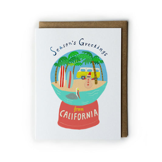 Holiday greeting card featuring a California snow globe with palm trees, beach, surfboard, vintage van, and snowman, set against a white card with a kraft paper envelope