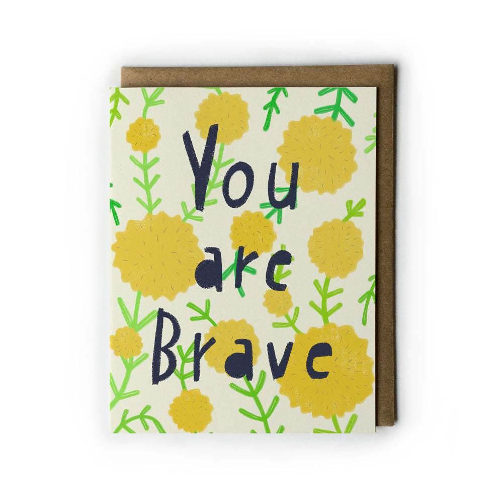 Greeting card with "You are Brave" text in blue, surrounded by yellow dandelion flowers and green stems on cream background, paired with brown kraft envelope