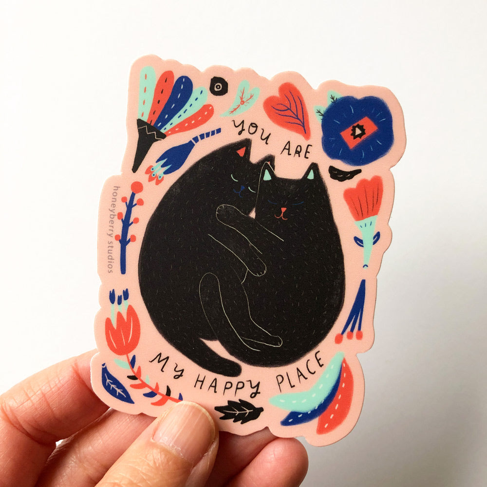 Colorful vinyl sticker featuring a black cat surrounded by floral and bird designs with text "You Are My Happy Place" on a pink background, held by fingertips