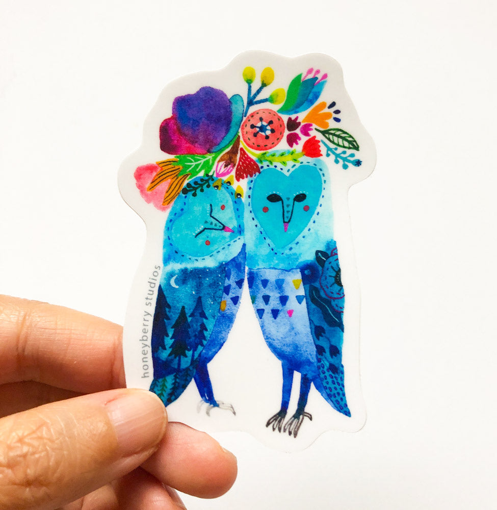 Colorful vinyl sticker featuring two blue barn owls cuddling with vibrant floral arrangement on their heads, held by a hand against white background
