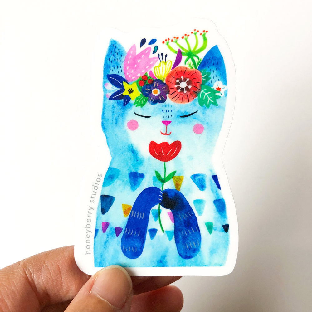 Colorful watercolor vinyl sticker of a cute blue cat with closed eyes, rosy cheeks, and a floral crown, holding a red flower. Hand shown holding the sticker for scale.