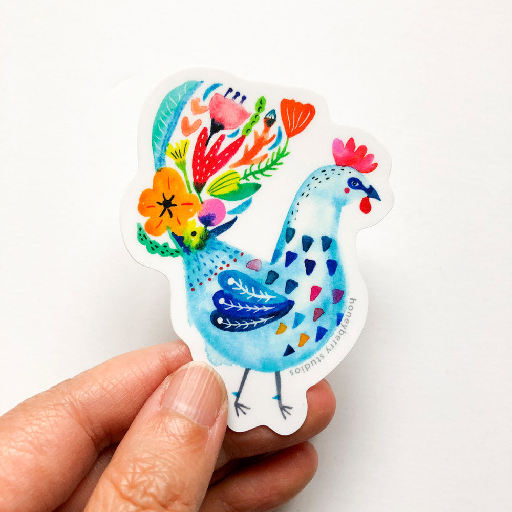 Colorful vinyl sticker of a blue rooster with floral tail feathers, featuring vibrant patterns and flowers in various hues, held between fingers against a white background
